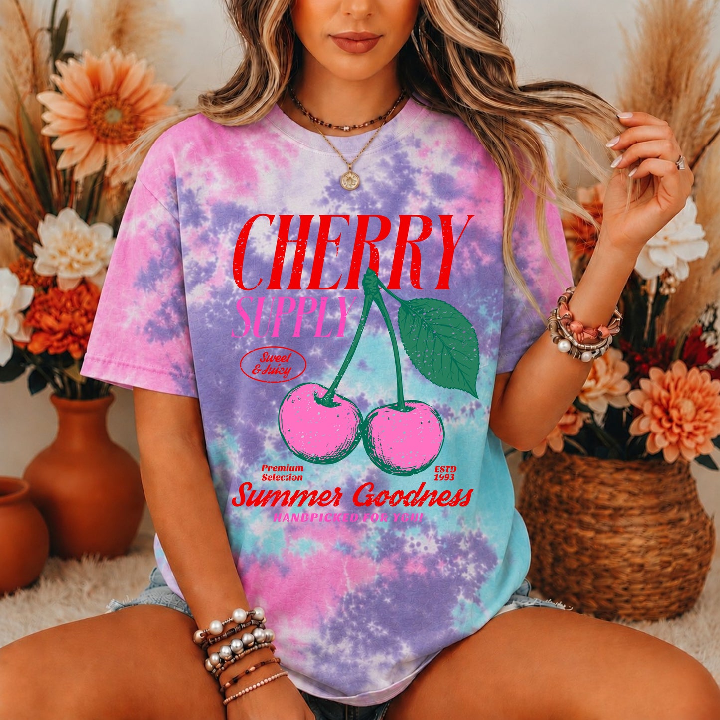 Person wearing a tie-dye t-shirt with cherry design and text, sitting in a decorative setting with flowers.