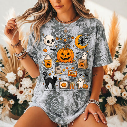 A woman wearing a gray acid-wash t-shirt with a colorful Halloween graphic print featuring pumpkins, ghosts, a black cat, and a crescent moon, posed in front of a floral background.